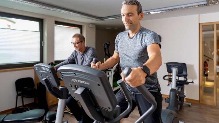 Zwei Männer trainieren in einem modernen Fitnessstudio auf Crosstrainern. Der Raum ist hell und gut ausgestattet.