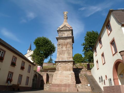 Ein historisches Denkmal steht an einer Straße, umgeben von typisch deutschen Gebäuden. Der Himmel ist blau und die Bäume sind grün.