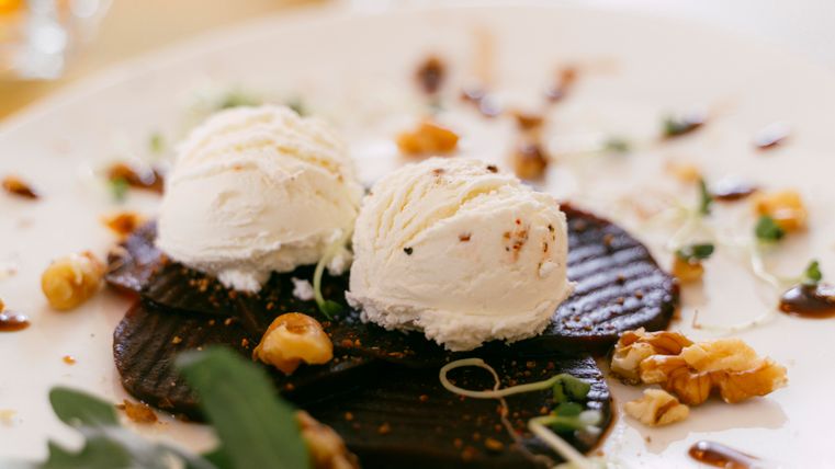 Ein Teller mit zwei Kugeln Vanilleeis auf einer Schokoladenbasis. Darauf sind Nüsse und eine dekorative Soße verteilt.