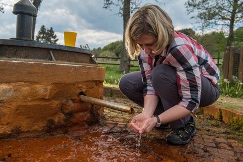 Eine Frau beugt sich über einen Brunnen und fängt Wasser mit ihren Händen auf. Im Hintergrund sind Bäume und ein orangefarbenes Trinkgefäß zu sehen.