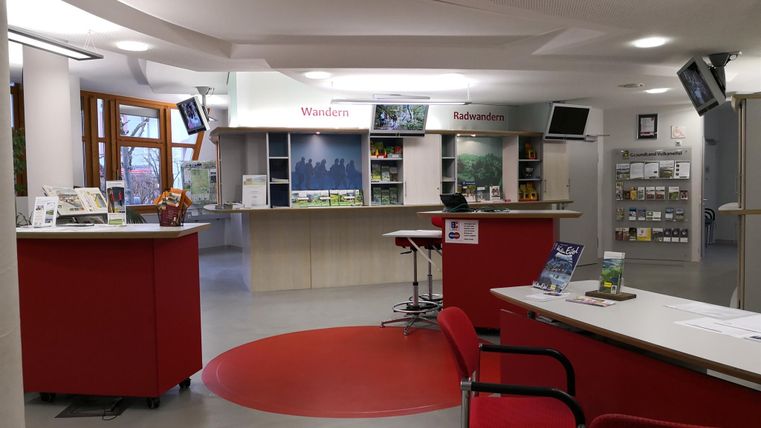 Een modern informatiecentrum met een receptiebalie en rode stoelen. Op planken liggen verschillende informatiematerialen over wandelen en vrijetijdsaanbiedingen.