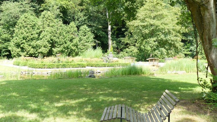 Ein ruhiger Garten mit einer Holzliege und grünen Bäumen. Im Hintergrund ist ein kleiner Teich sichtbar.