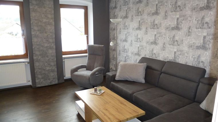 Ein modernes Wohnzimmer mit einem grauen Sofa und einem gemütlichen Sessel. Große Fenster sorgen für viel Tageslicht.