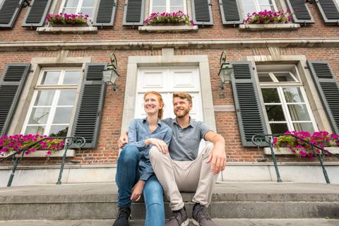 Ein Paar sitzt auf den Treppen vor einem schönen Gebäude mit Fensterläden. Die Fassade ist mit bunten Blumen geschmückt.
