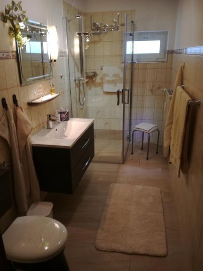 Ein modernes Badezimmer mit einer Dusche in Glasoptik und hellen Fliesen. Es gibt einen Waschbeckenunterschrank, Handtücher und eine weiche Badematte.