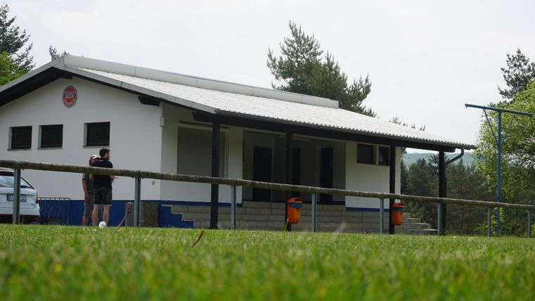 Ein weißes Gebäude mit Vordach und Treppe, umgeben von Rasen und Bäumen. Zwei Personen stehen davor.