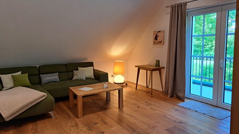 A cozy apartment with a green sofa and a wooden table. Large windows offer a view of the green surroundings.