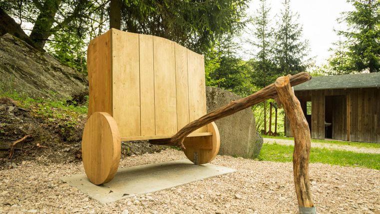 Holzskulptur eines Wagens auf einem Waldweg mit Bäumen im Hintergrund.