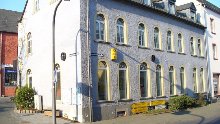 Historical building in Hillesheim with yellow post and tourism sign.
Street signs point to Koblenz and Aachen.