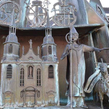 An artistic metal sculpture depicts a bishop with a staff and a church. In the background, further embellishments can be seen that evoke stories or traditions.