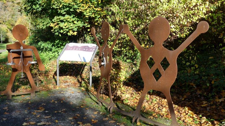 Three large, metallic figures stand along a path, surrounded by colorful autumn leaves. An information panel is located between the figures.