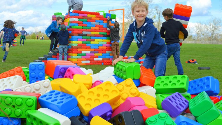 Ein Junge spielt mit bunten Bauklötzen im Freien. Im Hintergrund sind weitere Kinder beim Bauen einer großen Struktur zu sehen.