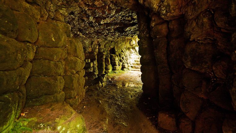 Een smalle, vochtige gang in een grot met een onregelmatige stenen muur. Het licht aan het einde van de gang zorgt voor een mystieke sfeer.