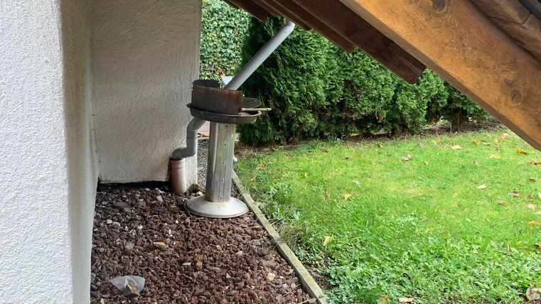 Eine Ecke eines Hauses mit einem geneigten Dach. Daneben steht ein Abflussrohr und im Hintergrund sieht man eine grüne Rasenfläche.