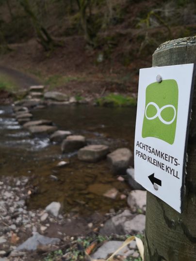 Ein Wegweiser mit dem Hinweis auf das "Achtsamkeits-Pfad Kleine Kyll" steht neben einem kleinen Fluss. Im Hintergrund sind Steine im Wasser zu sehen, die als Überquerungsmöglichkeiten dienen.