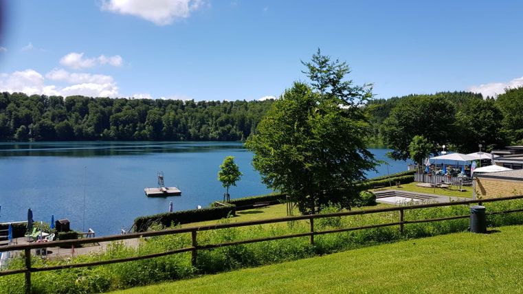 A tranquil lake surrounded by green trees and meadows. In the background, a pier and some lounge chairs can be seen.