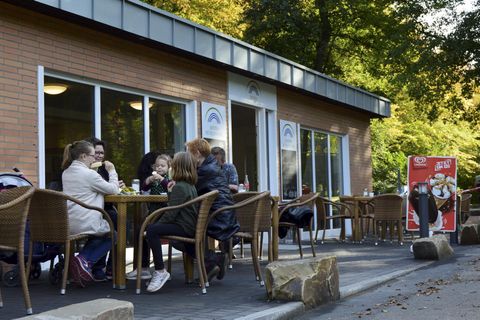 Ein Café mit mehreren Gästen, die draußen am Tisch sitzen. Im Hintergrund sind Bäume und ein Werbeplakat sichtbar.