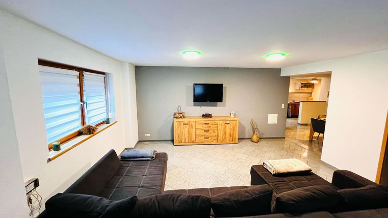 A modern living room with a black couch and a wooden cabinet. A television hangs on the wall and there are large windows with blinds.