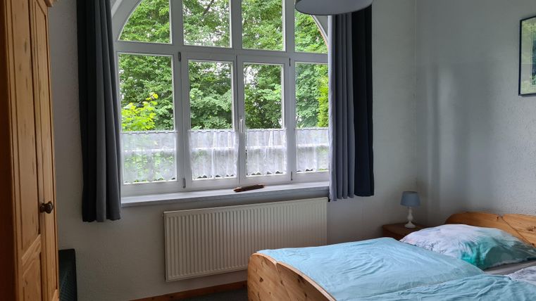 Ein helles Schlafzimmer mit einem großen Fenster und Blick ins Grüne. Es gibt ein Holzbett und einen Kleiderschrank.