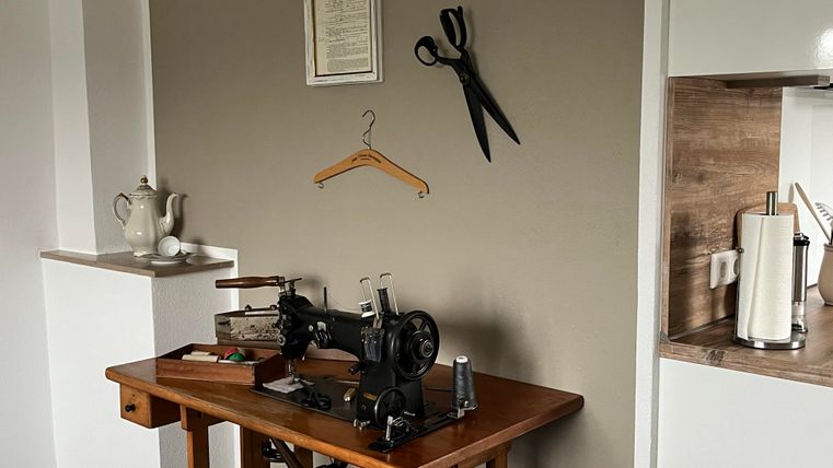 Ein Raum mit einer alten Nähmaschine auf einem Holztisch. An der Wand hängen eine Schere und ein Kleiderbügel.