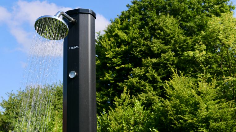 Een moderne buitendouche staat tegen een weelderig groene achtergrond. Het water stroomt stralend van de douchekop.