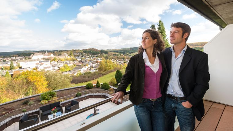 Ein Paar steht auf einem Balkon und schaut auf eine malerische Landschaft. Im Hintergrund sind Häuser und grüne Hügel unter einem bewölkten Himmel zu sehen.