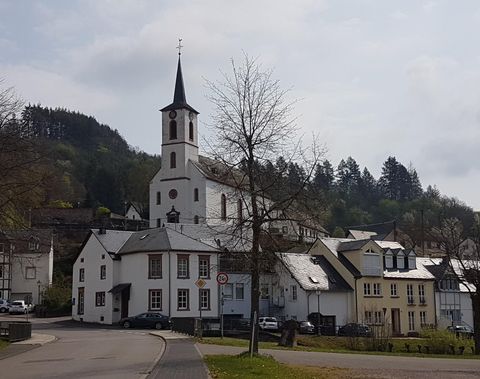 Een pittoresk dorpszicht met een kerk en verschillende historische gebouwen. Op de achtergrond zijn zacht glooiende heuvels en bomen te zien.