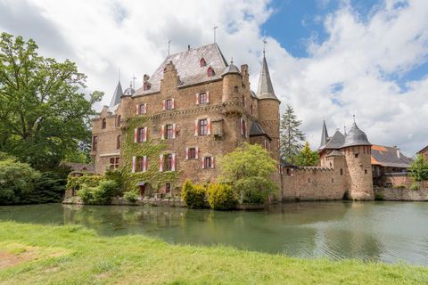 Ein schönes Schloss mit Türmen, umgeben von Bäumen und einem Teich. Der Himmel ist teilweise bewölkt und es ist eine ruhige Atmosphäre.