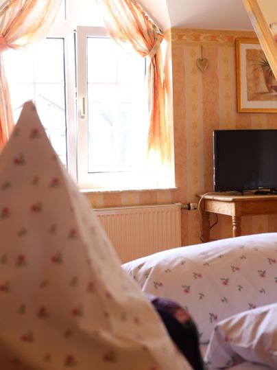 Ein gemütliches Zimmer mit hellen Wänden und einem großen Fenster. Im Hintergrund sind ein Fernseher und ein Holzstuhl zu sehen.