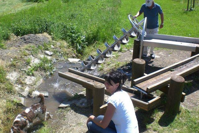 Eine Frau sitzt neben einem kleinen Wasserlauf und schaut zur Kamera. Im Hintergrund steht ein Mann an einer Wasserförderanlage und ein Hund spielt im Wasser.