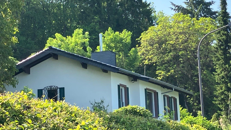 Ein helles Haus, umgeben von üppigem Grün und Bäumen. Der Himmel ist blau und klar.