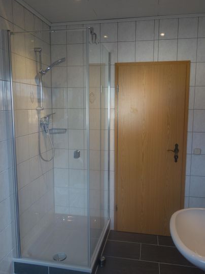 Een moderne badkamer met een glazen douchecel en een wastafel. De wanden zijn bekleed met witte tegels en er is een houten binnendeur zichtbaar.