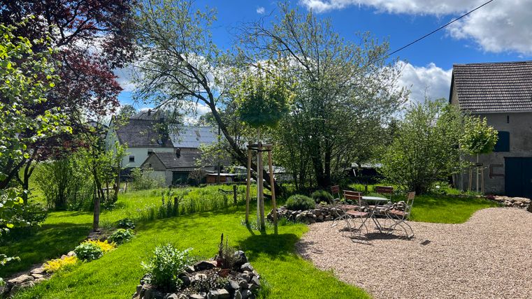 Eine grüne Gartenlandschaft mit blühenden Bäumen und einem hellen Himmel. In der Mitte sind Sitzgelegenheiten und ein kleiner Kiesweg.