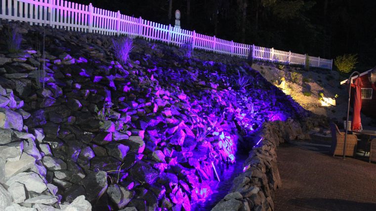 Ein beleuchteter Hang mit bunten Lichtern und Steinen. Im Hintergrund ist ein Zaun sichtbar.