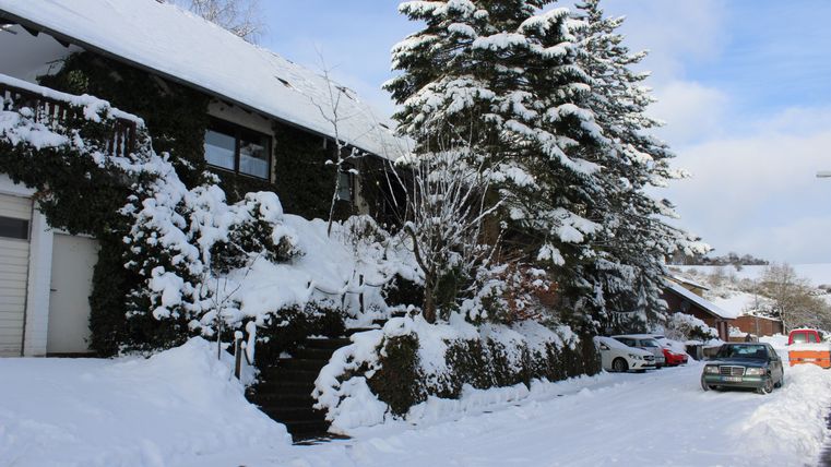 Ein winterliches Bild mit einem schneebedeckten Haus und hohen, verschneiten Tannenbäumen. Die Straße ist ebenfalls mit Schnee bedeckt und einige Autos sind sichtbar.