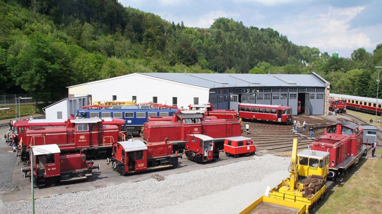 Een werkplaats voor treinen met veel rode locomotieven en goederenwagons. Op de achtergrond zijn groene bomen en een blauwe lucht te zien.