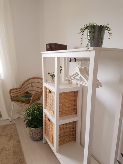 Een heldere ruimte met een witte kast die manden en kleerhangers bevat. In de hoek staat een gezellige rotanstoel en enkele planten zorgen voor een frisse sfeer.