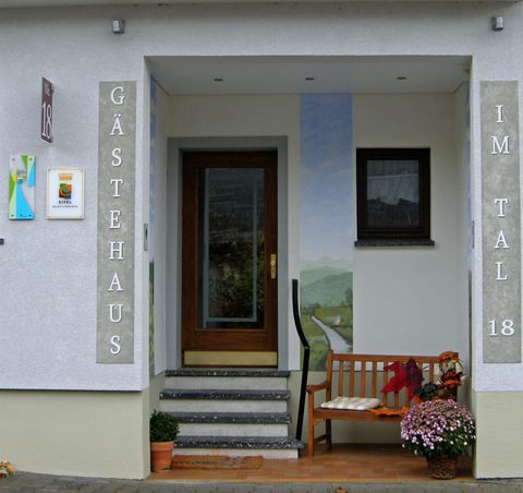 Een uitnodigende voordeur naar een gastenhuis met een gezellige bank en bloemen. De muur is gedeeltelijk versierd met een landschapsschilderij.
