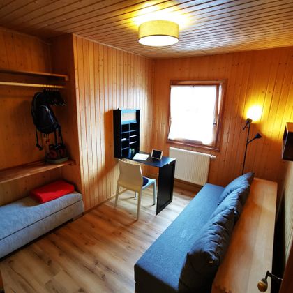 Een kleine kamer met houten bekleding. Er is een bed, een bureau en een stoel, evenals een raam dat natuurlijk licht binnenlaat.