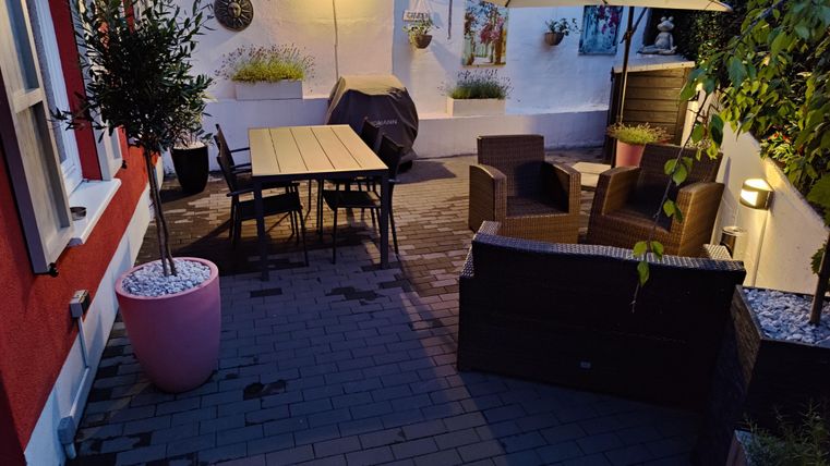 A cozy patio with a large table, chairs, and a sunshade. The surroundings are well-lit and there are some plants and decorations on the wall.