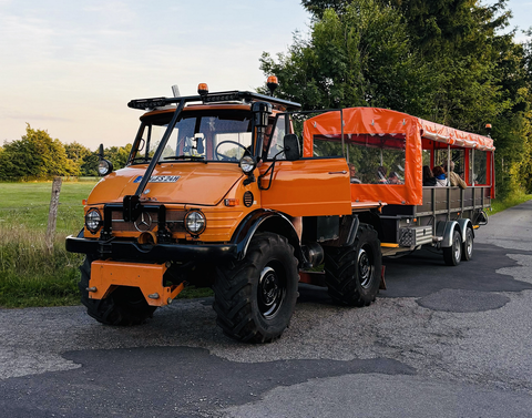 Ein orangefarbener Unimog mit einem Anhänger fährt auf einer ländlichen Straße. Im Anhänger sitzen Personen und die Umgebung ist grün und bewaldet.