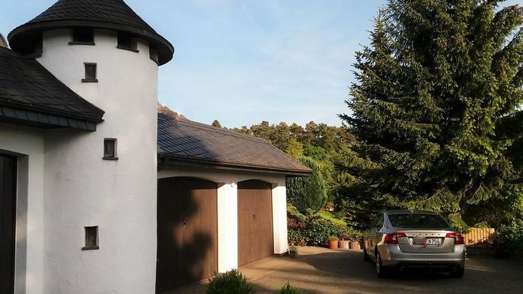 An elegant house with a round tower and a gray car in the driveway. Surrounded by trees and green scenery.