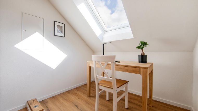 Ein heller Arbeitsraum mit einem Holzschreibtisch und einer weißen Stuhl. Ein kleines Pflänzchen und ein Dachfenster sorgen für eine freundliche Atmosphäre.
