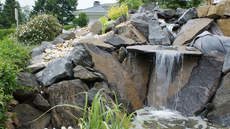 Ein schöner Wasserfall, der über große Steine fließt, umgeben von grünem Gras und Büschen. Im Hintergrund sind einige Bäume und ein Gebäude sichtbar.