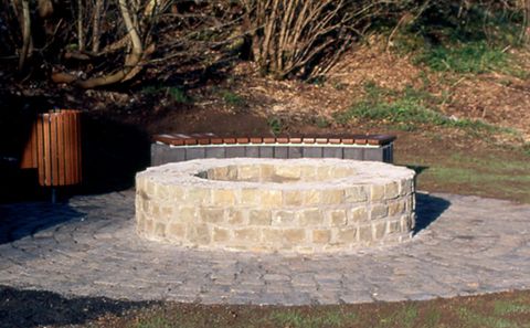 Runder, steinerner Brunnen mit Bank und Mülleimer im Hintergrund, umgeben von Bäumen.