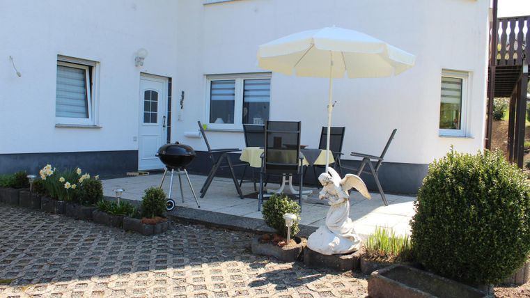 A cozy outdoor area with a table, chairs, and a sunshade. There is a grill and some plants as well as a statue.