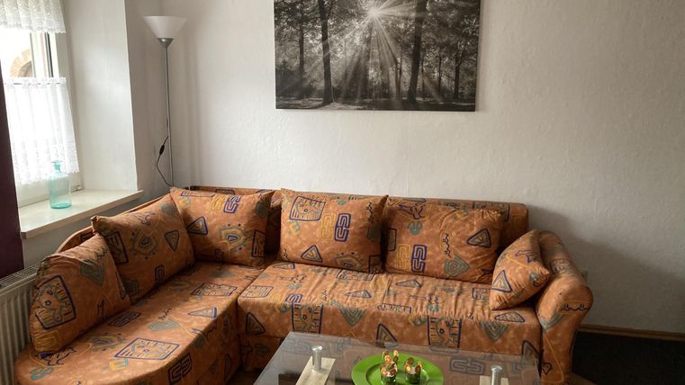 Een gezellige woonkamer met een oranje sofa-hoek en een glazen tafel. Aan de muur hangt een zwart-wit afbeelding van een bos.