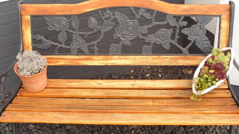 Eine Holzbank mit einem dekorativen Metallrücken. Neben der Bank stehen zwei Blumentöpfe mit Pflanzen.