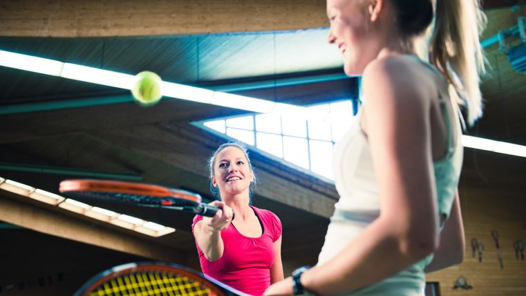 Twee vrouwen spelen tennis in een sporthal. Een speelster is klaar om de bal te raken, terwijl de andere glimlacht.