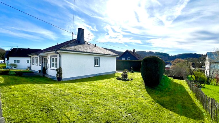 A small, white house with a well-maintained garden. In the background there are gentle hills and a blue sky with a few clouds.